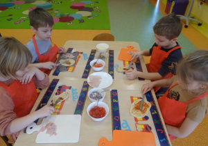 Czwórka dzieci smaruje sosem pomidorowym placek w kształcie koła przy użyciu noży.